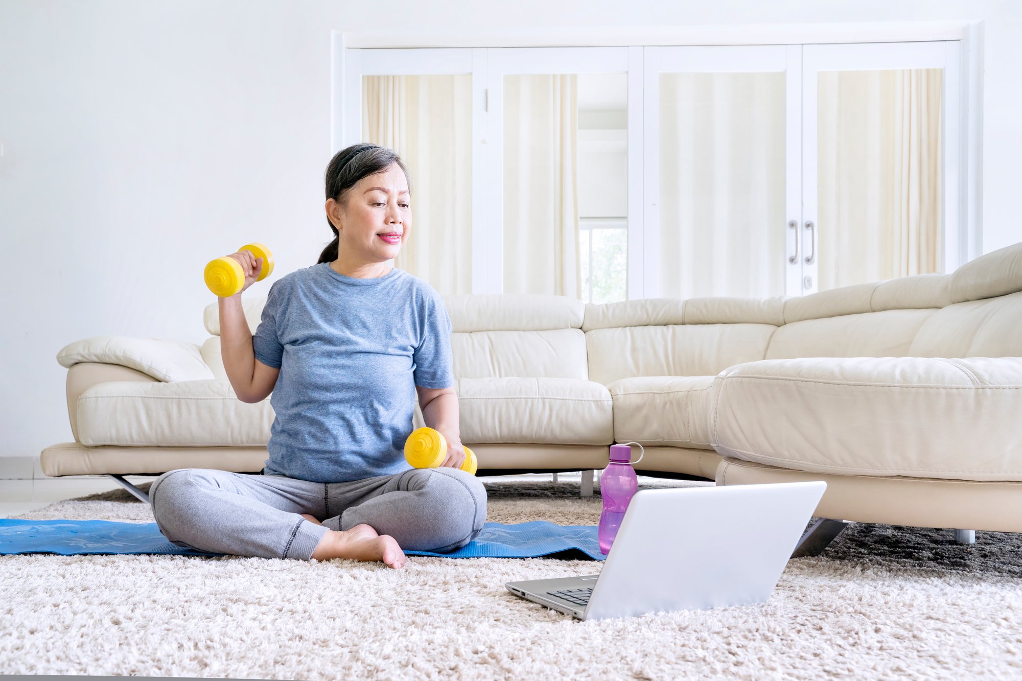 Asian women sitting on living floor watching a workout video with dumbbells in her hands, increasing her bone health and hormones through exercise.