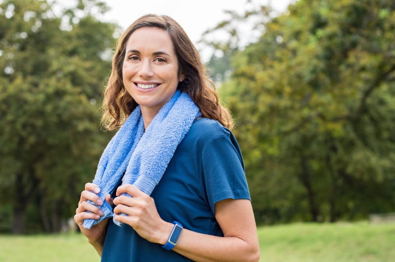 A woman holding a towel around her neck after working out in the park, her smile shows that exercise can help increase cellular energy and the metabolism.