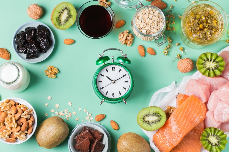 Green clock surrounded by fruit, nuts, meat, and grains on a mint background, showing that intermittent fasting improves bioenergetics.