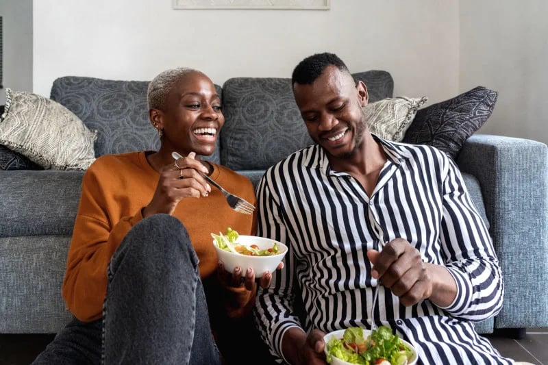 Cheerful young Black couple laughing while sitting on floor and eating salad in living room, happy to be utilizing functional medicine therapies to treat depression.
